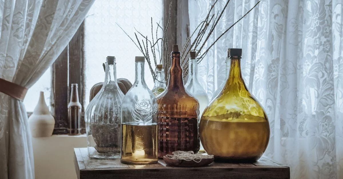 Présentation esthétique de diverses bouteilles en verre vintage sur une table en bois rustique près d'une fenêtre avec des rideaux en dentelle.