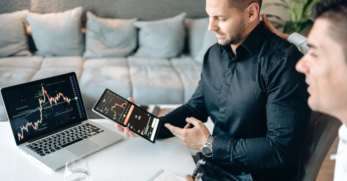 Deux hommes d'affaires discutent des données financières sur une tablette et un ordinateur portable, analysant des graphiques linéaires dans un bureau moderne.