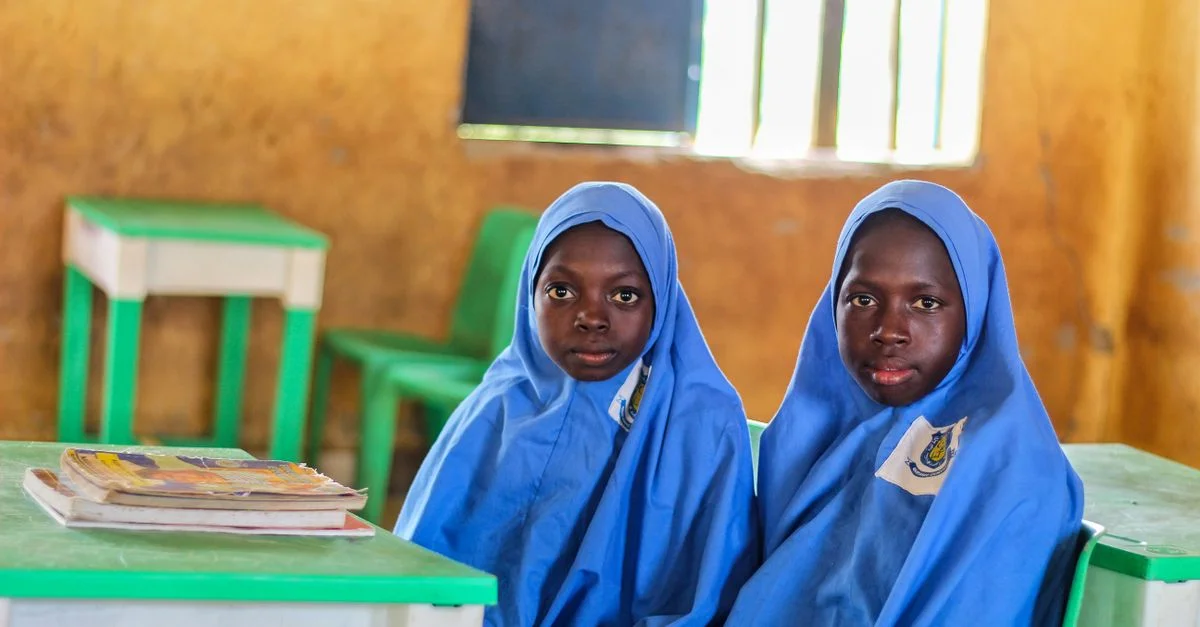 Deux jeunes élèves en hijabs bleus dans une salle de classe, assis près de livres et de bureaux.