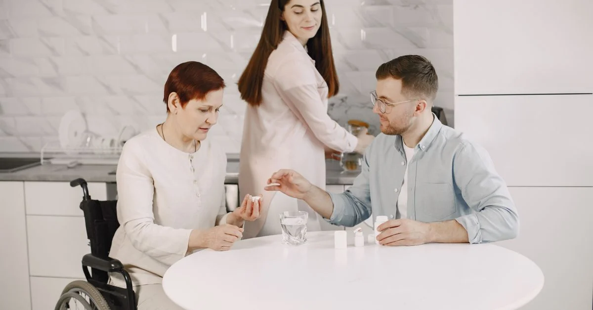 Femme âgée en fauteuil roulant recevant de l'aide pour ses médicaments d'un aidant dans une cuisine moderne.